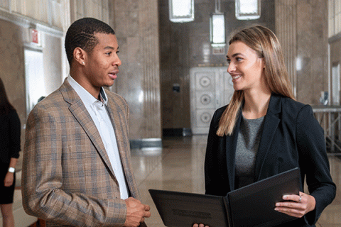 A white female student smiling in professional attire and holding a folder talking to a Black student in professional attire in the hallway of a building.